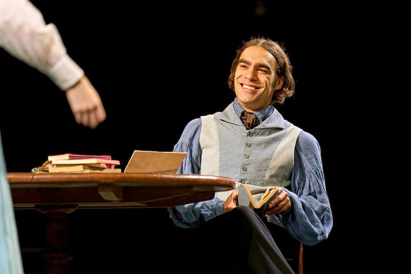 Seamus Dillane as Septimus, sitting and smiling at a table with books on it