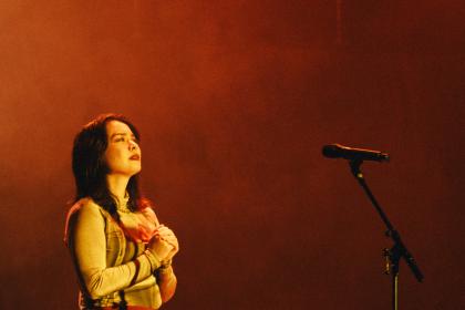 Mitski clutching her chest and looking upwards against a red background